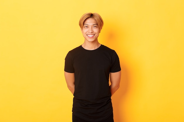 Retrato de amable chico asiático guapo con cabello rubio, sonriendo cortésmente, tomados de la mano detrás de la espalda, de pie pared amarilla