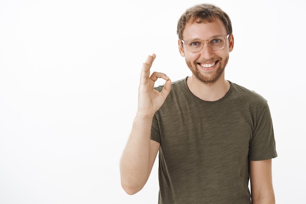 Retrato de amable guapo chico europeo servicial con gafas con barba mostrando gesto bien o bien y sonriendo asegurando que el trato con el cliente será firmado