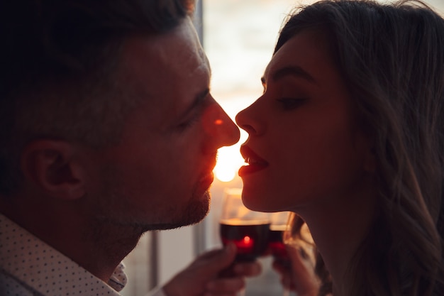 Retrato de amantes besándose al atardecer y sosteniendo un vaso con vino en casa