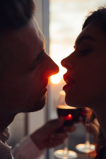 Retrato de amantes besándose al atardecer y sosteniendo un vaso con vino en casa