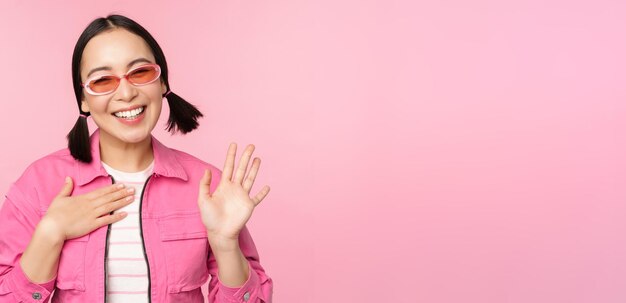 Retrato de una amigable adolescente asiática con gafas de sol que dice hola agita su mano y sonríe y te saluda