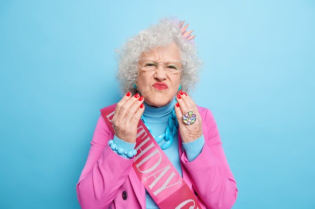 Retrato de anciana con estilo con el pelo gris y rizado pucheros labios pintados de rojo hace griace infeliz viste ropa de moda y una pequeña corona en la cabeza celebra birhday quiere ser siempre joven