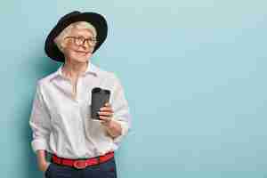 Foto gratuita retrato de anciana feliz que está en pensión, se reúne con ex colegas, sostiene café para llevar, viste elegante camisa blanca, pantalón con cinturón rojo, mantiene la mano en el bolsillo. ocio, jubilación