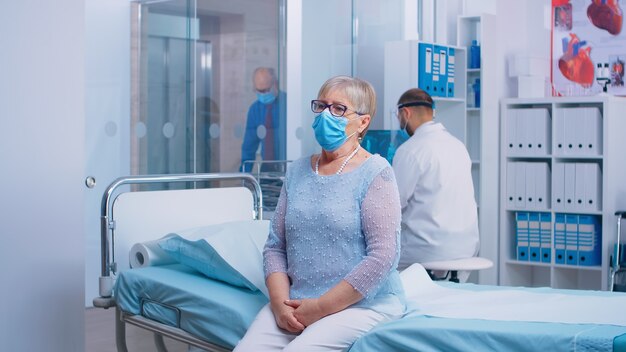 Retrato de anciana sentada en la cama del hospital durante la crisis de COVID-19. Médico y pacientes con máscara protectora y equipo en cinic privado moderno. Sistema de salud después del coronavirus