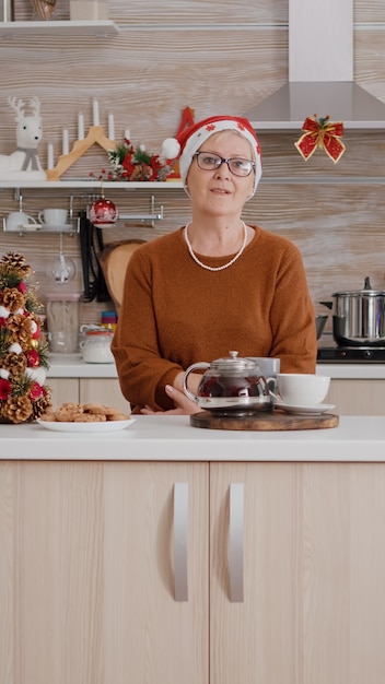 Retrato de anciana con sombrero de Navidad celebrando la temporada navideña en cocina decorada