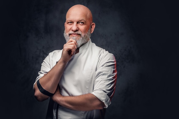 Retrato de un anciano chef pensativo vestido con uniforme posando sobre un fondo oscuro.