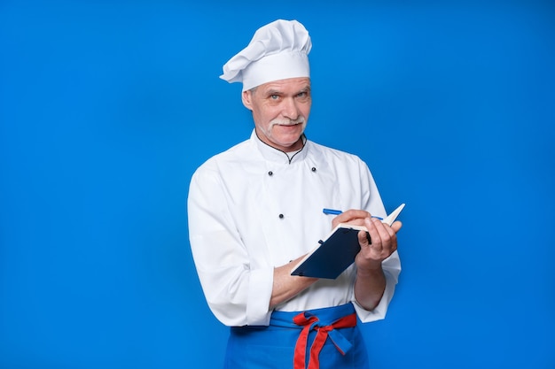 Retrato de anciano chef principal aislado en la pared azul, con su bloc de notas, escribe una nueva receta.