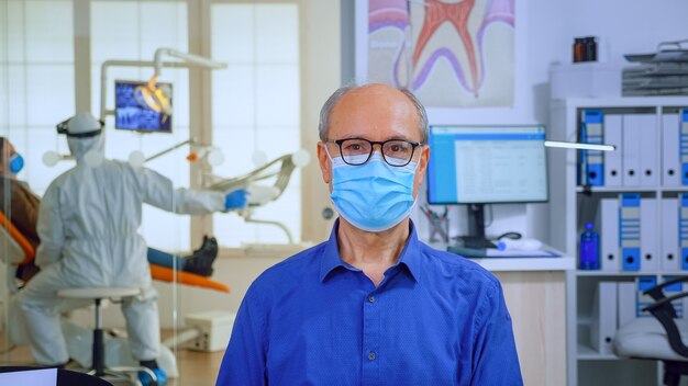 Retrato de anciano en consultorio dental mirando a cámara con mascarilla sentado en una silla en la sala de espera de la clínica estomatológica. Concepto de nueva visita normal al dentista en caso de brote de coronavirus.