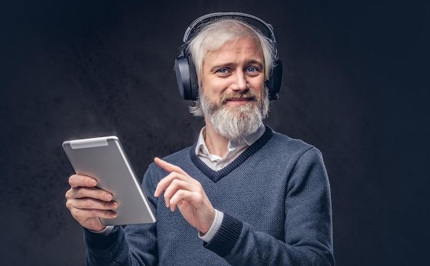 Retrato de un apuesto anciano usando una tableta con auriculares en un estudio sobre un fondo oscuro.