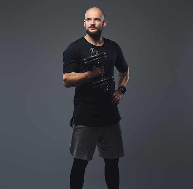 Retrato de un apuesto atleta barbudo con una camiseta negra, posando con pesas en un estudio. Aislado en un fondo gris.