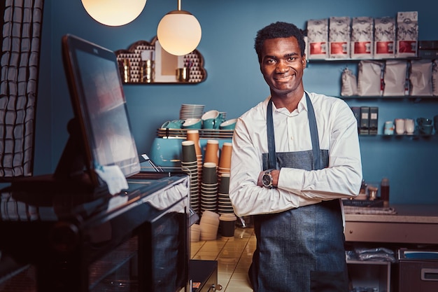 Foto gratuita retrato de un apuesto barista africano parado con los brazos cruzados en el mostrador de una cafetería de moda.