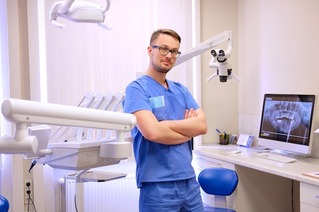 Retrato de un apuesto dentista con uniforme azul, de pie en una clínica de dentista. Mirando a la cámara.