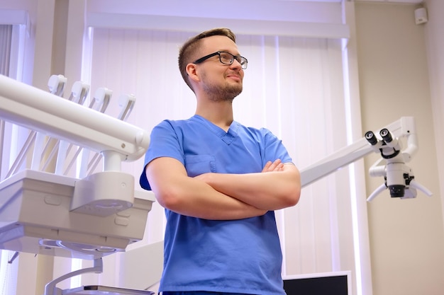 Retrato de un apuesto dentista con uniforme azul, de pie en una clínica de dentista.