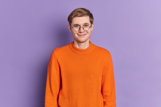Retrato de un apuesto estudiante europeo tiene una sonrisa suave en la cara feliz de escuchar agradables puestos de noticias encantado lleva gafas redondas jersey naranja