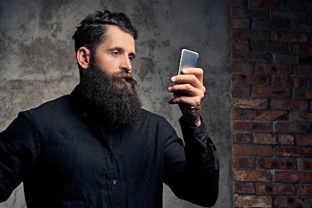 Retrato de un apuesto hipster barbudo vestido con una camisa negra, sostiene un teléfono inteligente, parado en un estudio. Aislado en un fondo oscuro.