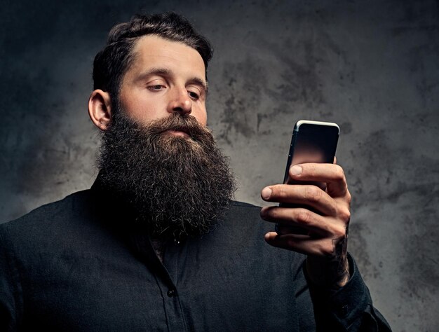 Retrato de un apuesto hipster barbudo vestido con una camisa negra, usando un smartphone, parado en un estudio. Aislado en un fondo oscuro.