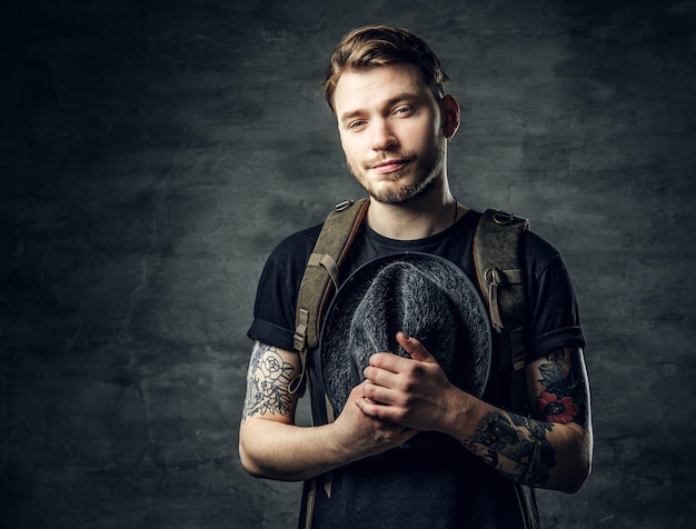 Foto gratuita retrato de un apuesto hipster viajero tatuado sostiene un sombrero de tweed en los brazos y una mochila en la espalda.