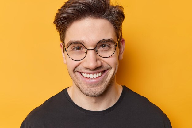 Retrato de un apuesto hombre adulto alegre que sonríe con dientes mira directamente a la cámara a través de gafas transparentes de buen humor vestido casualmente aislado sobre un fondo amarillo Emociones humanas