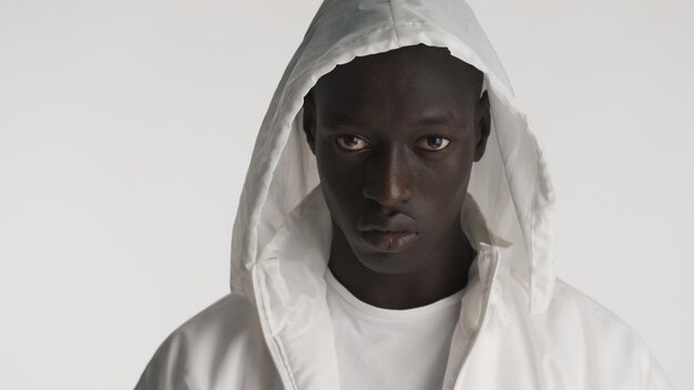 Retrato de un apuesto hombre afroamericano vestido con una chaqueta blanca con capucha posando en la cámara con una mirada seria aislada de fondo blanco