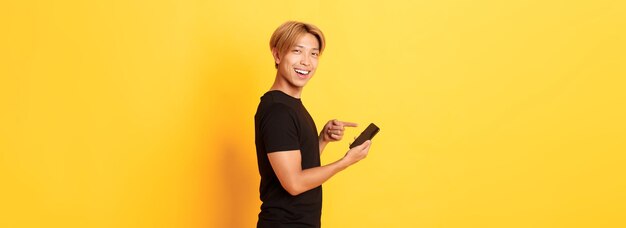 Retrato de un apuesto hombre asiático sonriente complacido de perfil y señalando con el dedo al teléfono inteligente