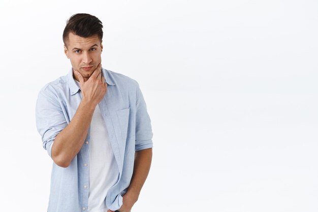 Retrato de un apuesto hombre macho con un corte de pelo elegante que toca la mandíbula limpia y afeitada y mira una cámara pensativa pensando en considerar y tomar una decisión ponderando la decisión de fondo blanco