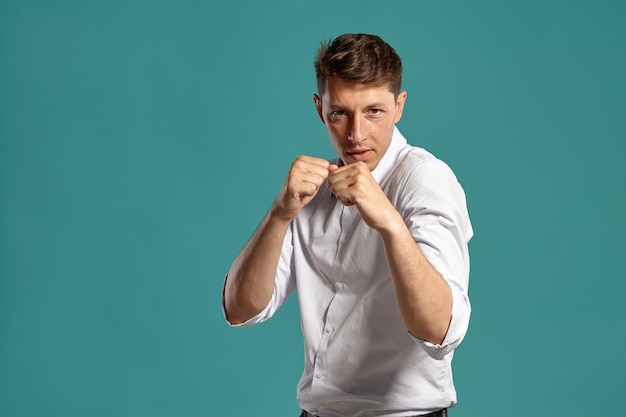 Foto gratuita retrato de un apuesto hombre moreno con ojos marrones, vestido con una camisa blanca clásica. está actuando como si estuviera peleando mientras posa en un estudio contra un fondo azul. concepto de gesticulación y s