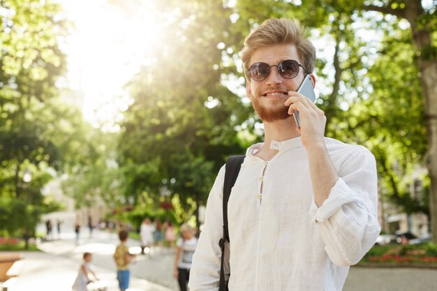 Retrato de apuesto hombre pelirrojo en camisa blanca y gafas de sol sonriendo, hablando por teléfono en el camino en la reunión de trabajo en la cafetería.