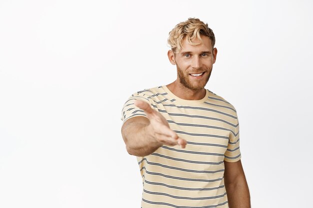 Retrato de un apuesto hombre rubio extendiendo la mano para un apretón de manos saludándote de pie en camiseta sobre fondo blanco