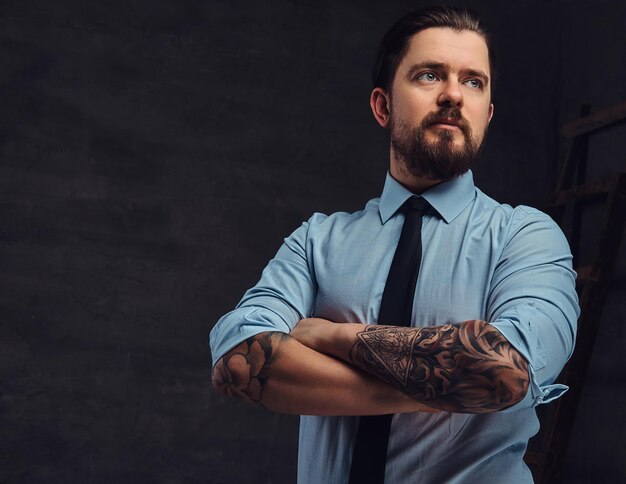 Retrato de un apuesto hombre tatuado de mediana edad con barba y peinado vestido con una camisa azul y corbata, posa en un estudio con los brazos cruzados. Aislado en un fondo oscuro texturizado en estudio.