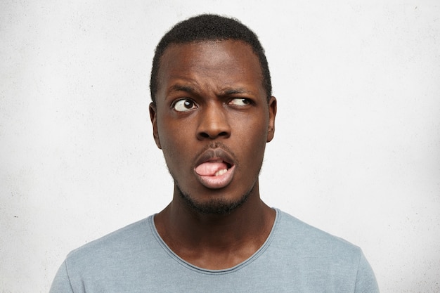 Retrato de apuesto joven africano vestido con camiseta gris mirando hacia arriba, sacando la lengua