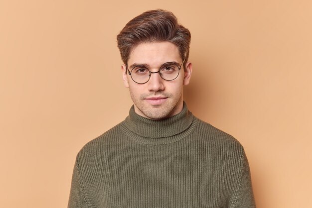 El retrato de un apuesto joven europeo mira directamente a la cámara con una expresión seria, se siente seguro de sí mismo, usa gafas redondas transparentes y un suéter casual aislado sobre fondo beige.