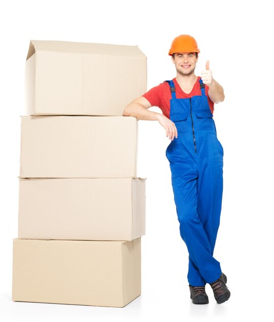 Retrato de apuesto joven repartidor con cajas de papel mostrando el pulgar hacia arriba signo aislado en blanco