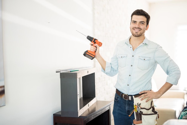 Foto gratuita retrato de un apuesto manitas hispano que sostiene un taladro eléctrico y sonríe mientras trabaja en una casa