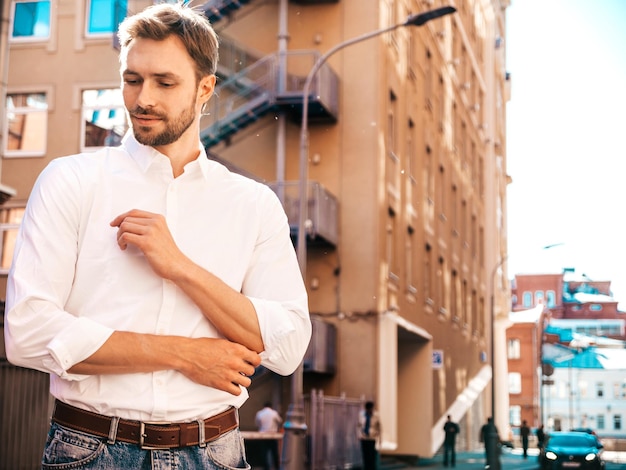 Retrato de un apuesto modelo lambersexual hipster elegante y confiado Hombre moderno vestido con camisa blanca Moda masculina pensativa posando en el fondo de la calle Al aire libre al atardecer