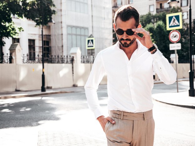 Retrato de un apuesto modelo lambersexual hipster elegante y confiado Hombre moderno vestido con camisa blanca y pantalones Hombre de moda posando en el fondo de la calle en la ciudad de Europa al atardecer Con gafas de sol