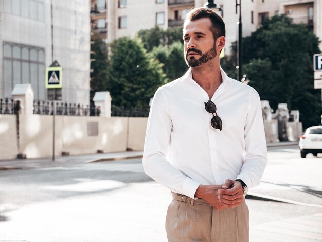 Retrato de un apuesto modelo lambersexual hipster elegante y confiado Hombre moderno vestido con camisa blanca y pantalones Hombre de moda posando en el fondo de la calle en la ciudad de Europa al atardecer Con gafas de sol