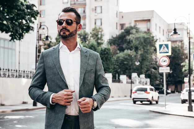 Retrato de un apuesto modelo lambersexual hipster elegante y confiado Hombre moderno vestido con traje elegante Hombre de moda posando en el fondo de la calle en la ciudad de Europa al atardecer Con gafas de sol