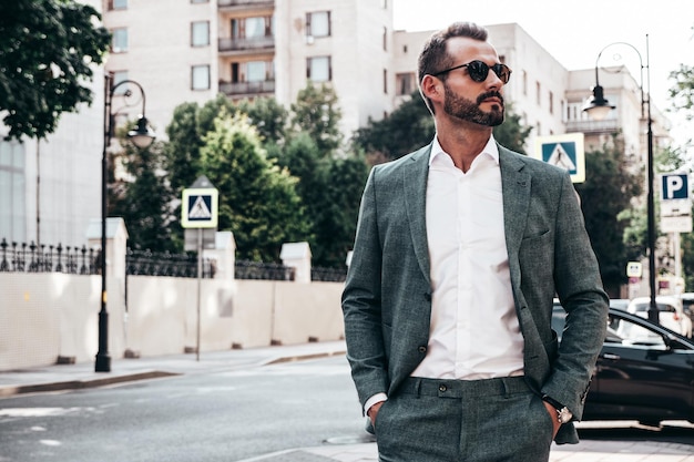 Retrato de un apuesto modelo lambersexual hipster elegante y confiado Hombre moderno vestido con traje elegante Hombre de moda posando en el fondo de la calle en la ciudad de Europa al atardecer Con gafas de sol