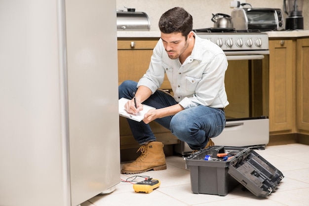 Retrato de un apuesto técnico masculino haciendo un informe de trabajo sobre una nevera rota en la cocina de una casa