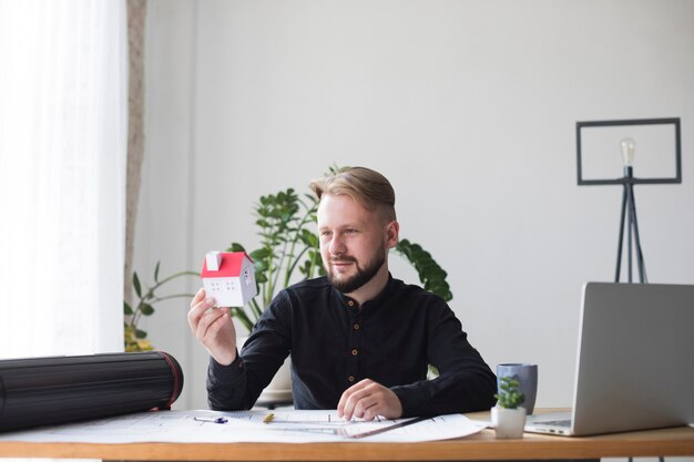 Retrato de una arquitectura masculina joven que sostiene el modelo de la casa mientras que se sienta en oficina