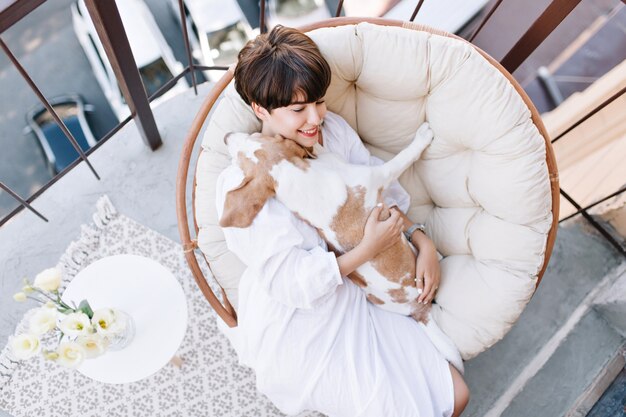 Retrato desde arriba de la alegre señorita descansando en el balcón después de la ducha y acariciando suavemente la mascota. Adorable niña pasar tiempo en la terraza con beagle disfrutando el fin de semana