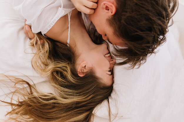 Retrato de arriba de dos jóvenes enamorados que se divierten juntos en la acogedora cama por la mañana. Chica morena de pelo largo riendo mientras su novio la abraza descansando al lado