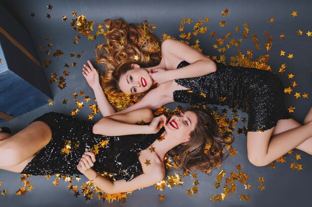 Retrato de arriba de dos niñas alegres en confeti dorado. Señora de pelo largo en vestido negro divirtiéndose con hermana morena en fiesta de año nuevo.