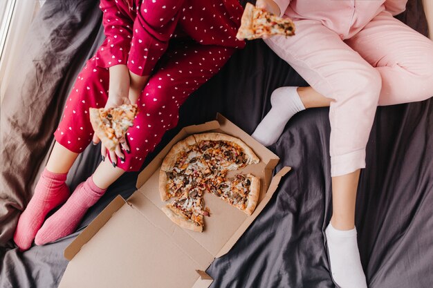 Retrato de arriba de dos niñas en pijama sentada en la cama con comida rápida italiana. Modelos femeninos perezosos comiendo pizza en la hoja oscura.