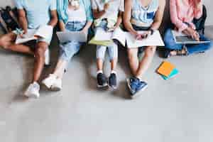 Foto gratuita retrato de arriba de jóvenes con portátiles y teléfonos inteligentes, sentados juntos en el suelo. estudiantes escribiendo conferencias sosteniendo libros de texto de rodillas.