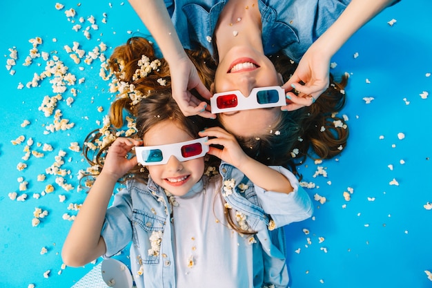 Retrato desde arriba linda madre e hija poniendo cabeza a cabeza aislado en piso azul. Usar lentes 3D, cabello largo moreno, divertirse con palomitas de maíz, mejores fines de semana, tiempo libre con la familia