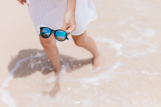 Retrato de arriba de la mujer de pie en el agua en la costa. Foto exterior de refinada mujer bronceada con gafas de sol brillantes en la mano.