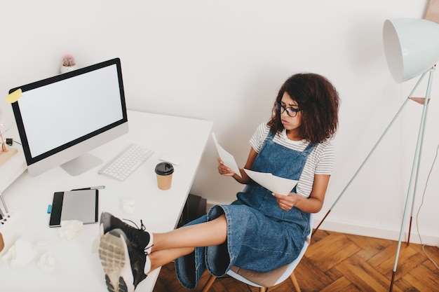 Retrato desde arriba de la ocupada mujer joven con gafas sentado con las piernas en la mesa junto a la computadora y la tableta