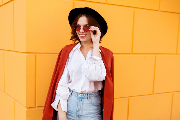 Retrato ascendente cercano del exterior de la hembra de pelo corto morena feliz que presenta sobre la pared amarilla. Sombrero de moda, lentes rosas, blusa blanca y jeans.