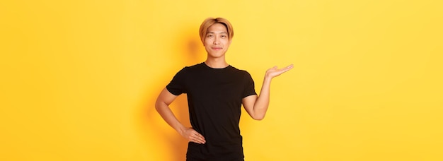 Retrato de un asiático sonriente feliz y orgulloso sosteniendo algo en la mano sobre un fondo amarillo